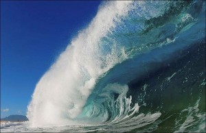 A large ocean wave curling with white foam under a blue sky.